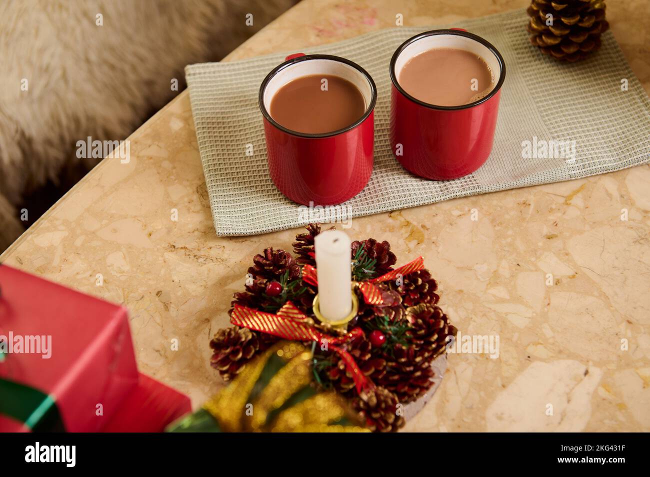 Nahaufnahme von roten Tassen mit heißem Schokoladengetränk auf grüner Serviette, neben weihnachtlich beleuchteter Kerze und Geschenken auf Marmortisch Stockfoto