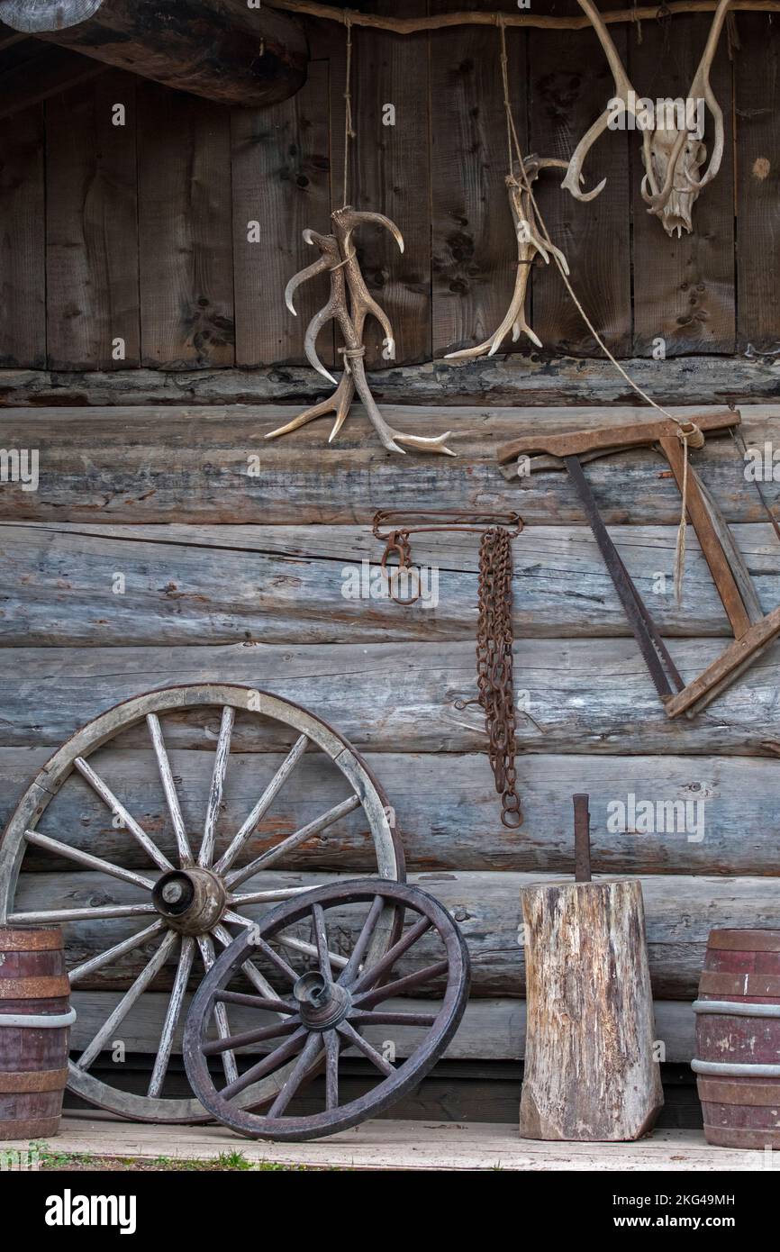 Alte Holzfässer, Wagenräder und rostige Ketten, Hirschgeweihe und antike Holzsägen, die draußen an der Wand der Trapper-Blockhütte hängen Stockfoto