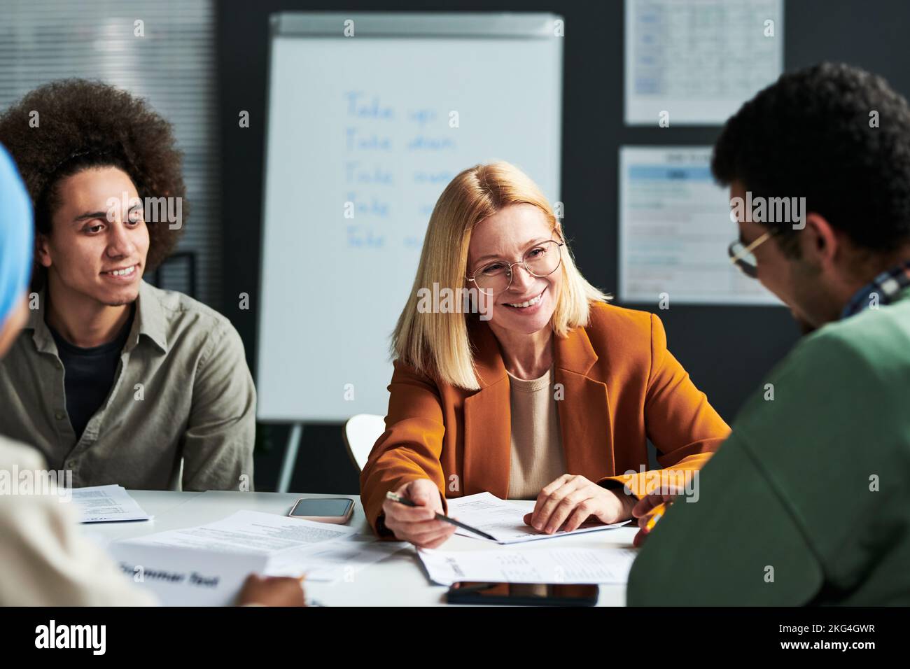 Glückliche reife blonde Linguistik-Lehrerin, die einem Schüler etwas erklärt, während sie ihn anschaut und auf ein Dokument zeigt Stockfoto