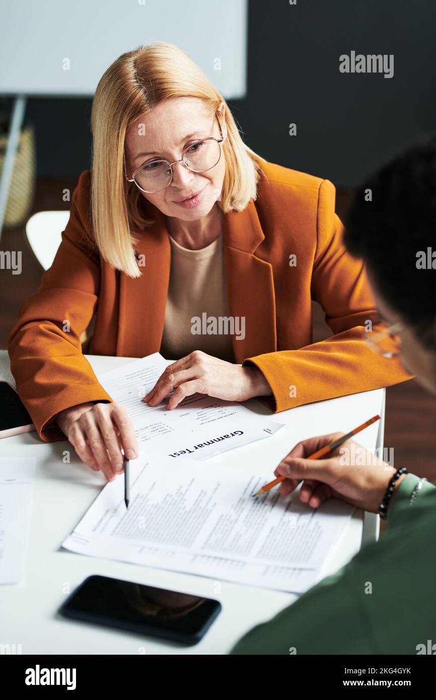 Selbstbewusste blonde reife Linguistiklehrerin, die jungen Mann unterrichtet, während sie auf eines der Dokumente im Unterricht in der Highschool zeigt Stockfoto