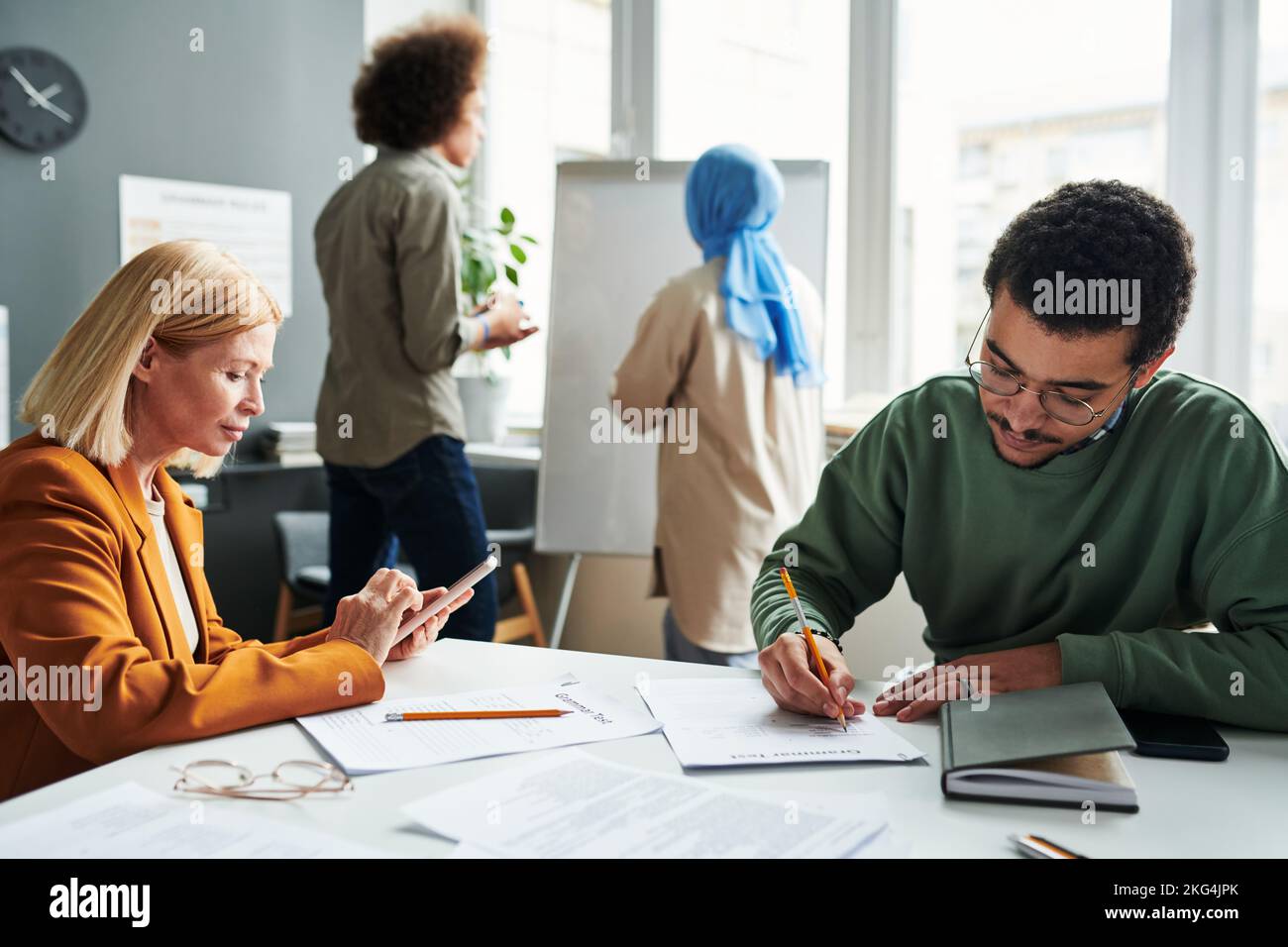 Reifer blonder Lehrer und männlicher Schüler aus dem Nahen Osten, der individuelle Arbeit leistet, während er am Tisch gegen zwei Lernende am Whiteboard sitzt Stockfoto