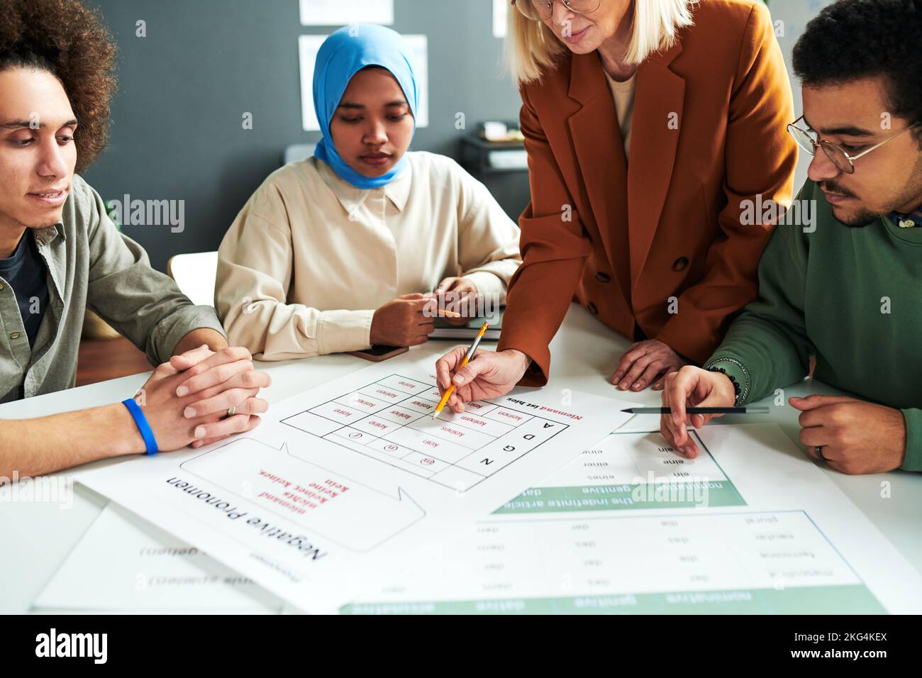Reifer blonder Lehrer, der sich über Dokument mit Pronomen beugt, während er der Gruppe interkultureller Schüler neues Thema erklärt Stockfoto