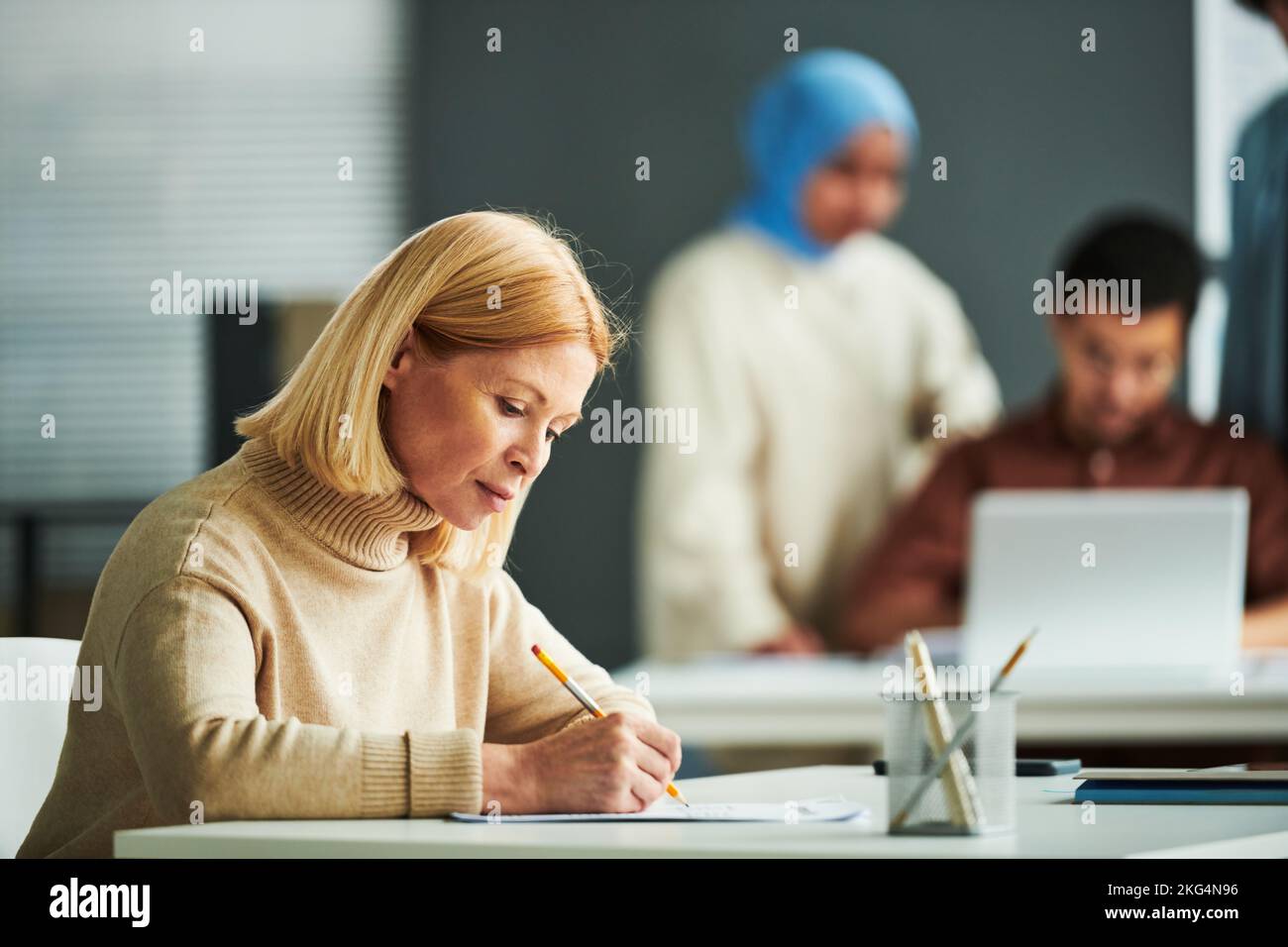 Seriöse reife blonde Lehrerin in beigefarbenem Pullover beim Grammatiktest, während sie vor der Kamera vor dem Schreibtisch gegen zwei Schüler sitzt Stockfoto