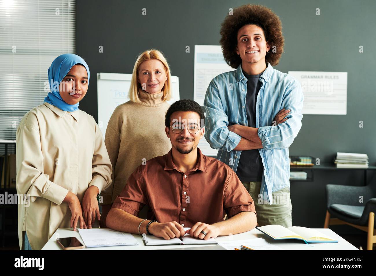 Eine Gruppe multikultureller Schüler und reifer blonder Lehrer in lässiger Kleidung, die vor der Kamera mit Whiteboard hinter dem Schreibtisch stehen Stockfoto
