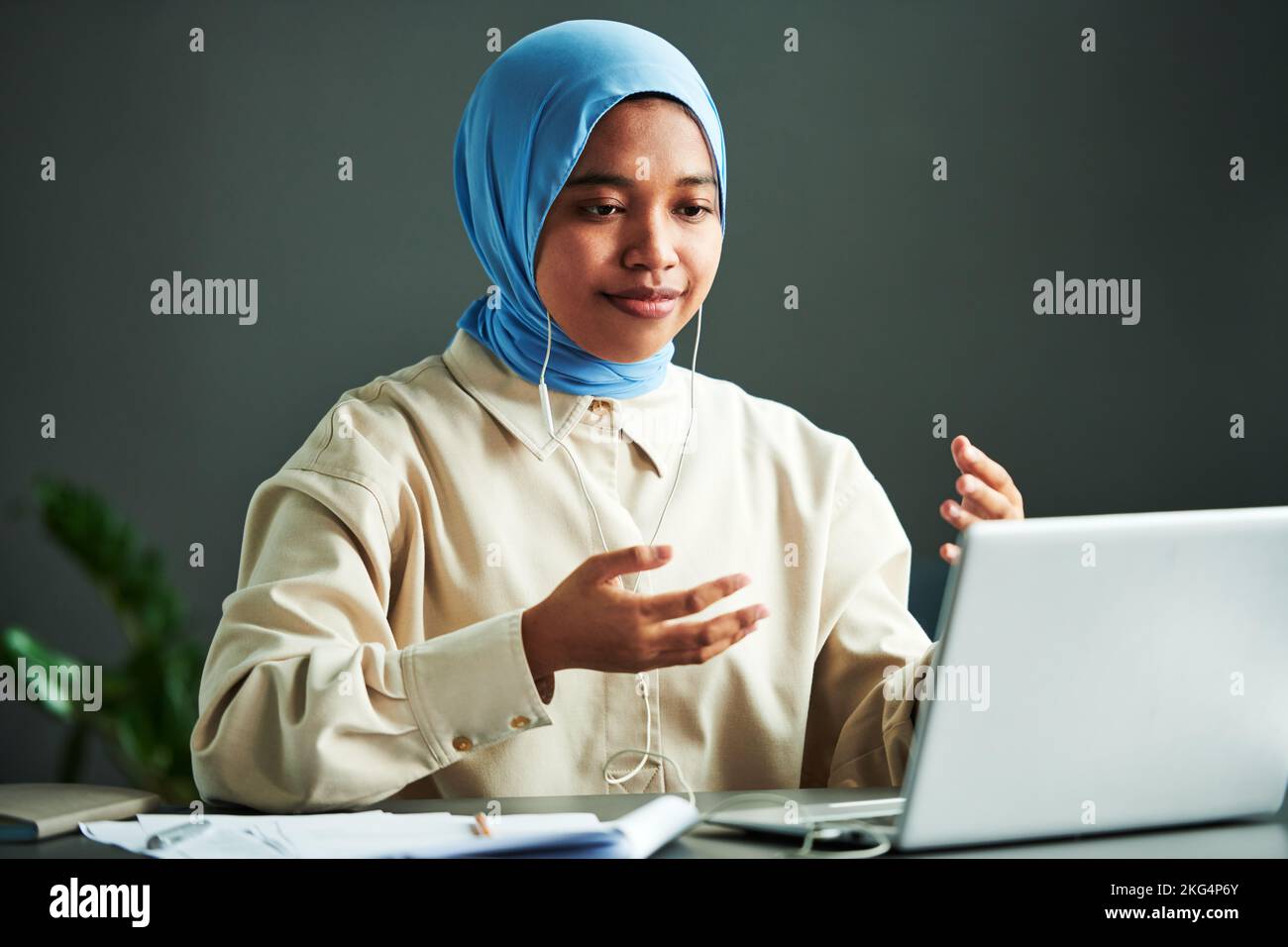 Junge selbstbewusste muslimische Frau in Casualwear und blauem Hijab, die während des Online-Unterrichts vor dem Laptop etwas erklärt Stockfoto