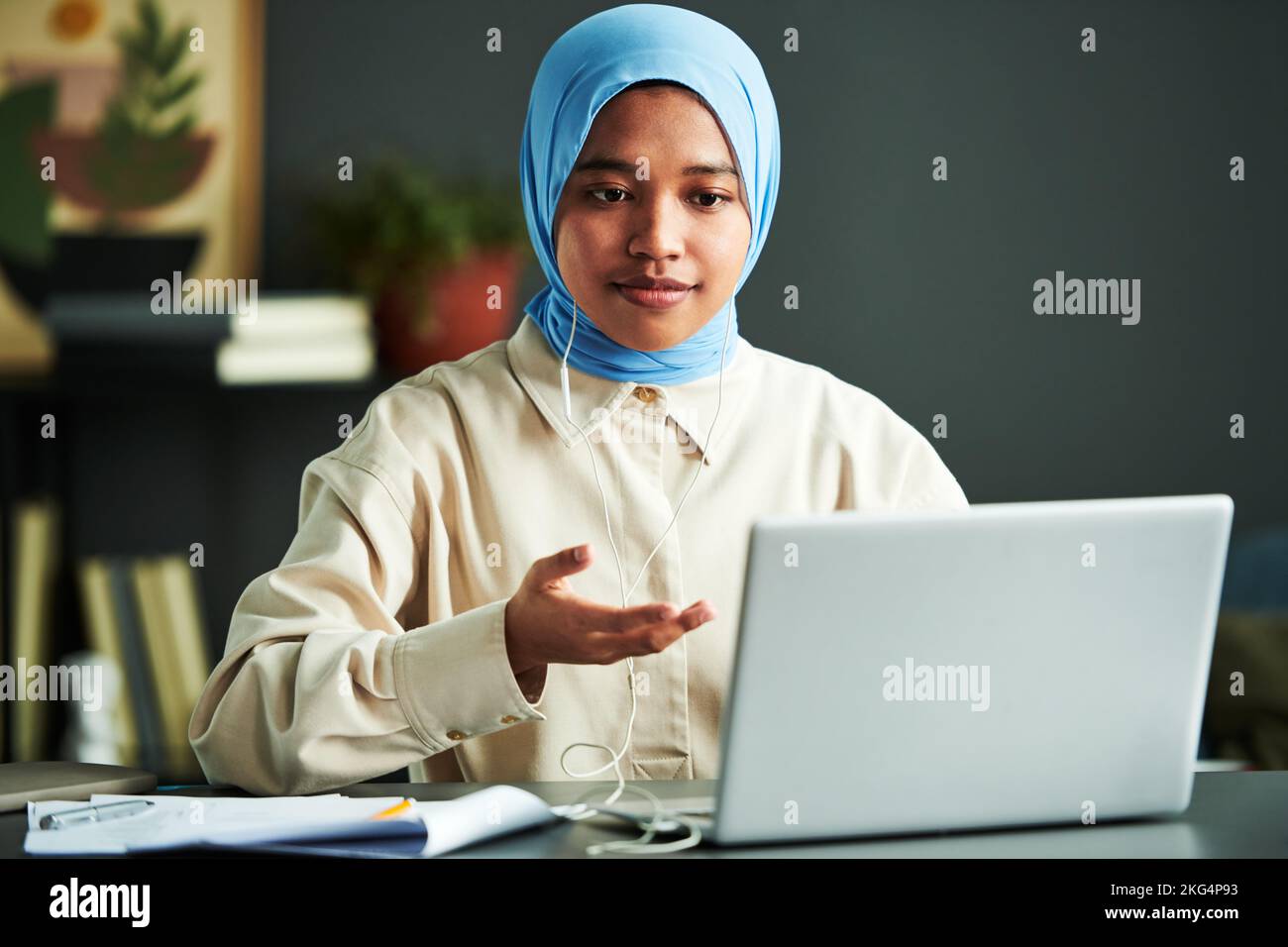 Junge selbstbewusste muslimische Frau in Hijab, die am Webinar teilnimmt, während sie zu Hause vor dem Laptop am Schreibtisch sitzt und etwas erklärt Stockfoto