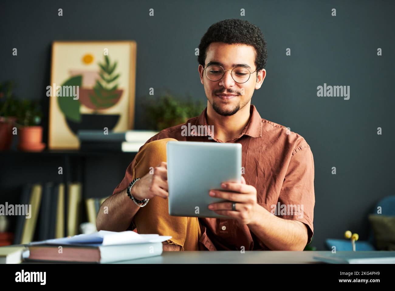 Junger männlicher Student aus dem Nahen Osten schaut sich Online-Informationen auf dem Tablet-Bildschirm an, während er mit Büchern am Arbeitsplatz sitzt Stockfoto