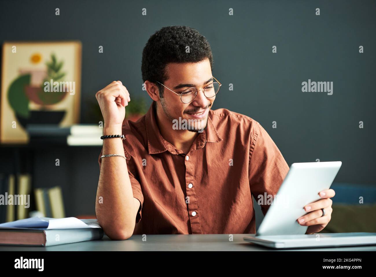 Junger lächelnder männlicher Student des Online-Studiengangs beim Betrachten des Tablet-Bildschirms beim Sitzen am Schreibtisch und beim Betrachten der Videostunde Stockfoto
