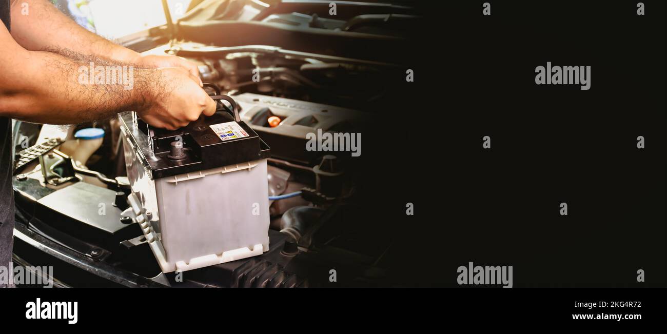Ein Automechaniker trägt eine Ersatz-Autobatterie für die elektrische Wartung des Autos, Panorama-Banner mit Kopierraum auf schwarzem Hintergrund Stockfoto