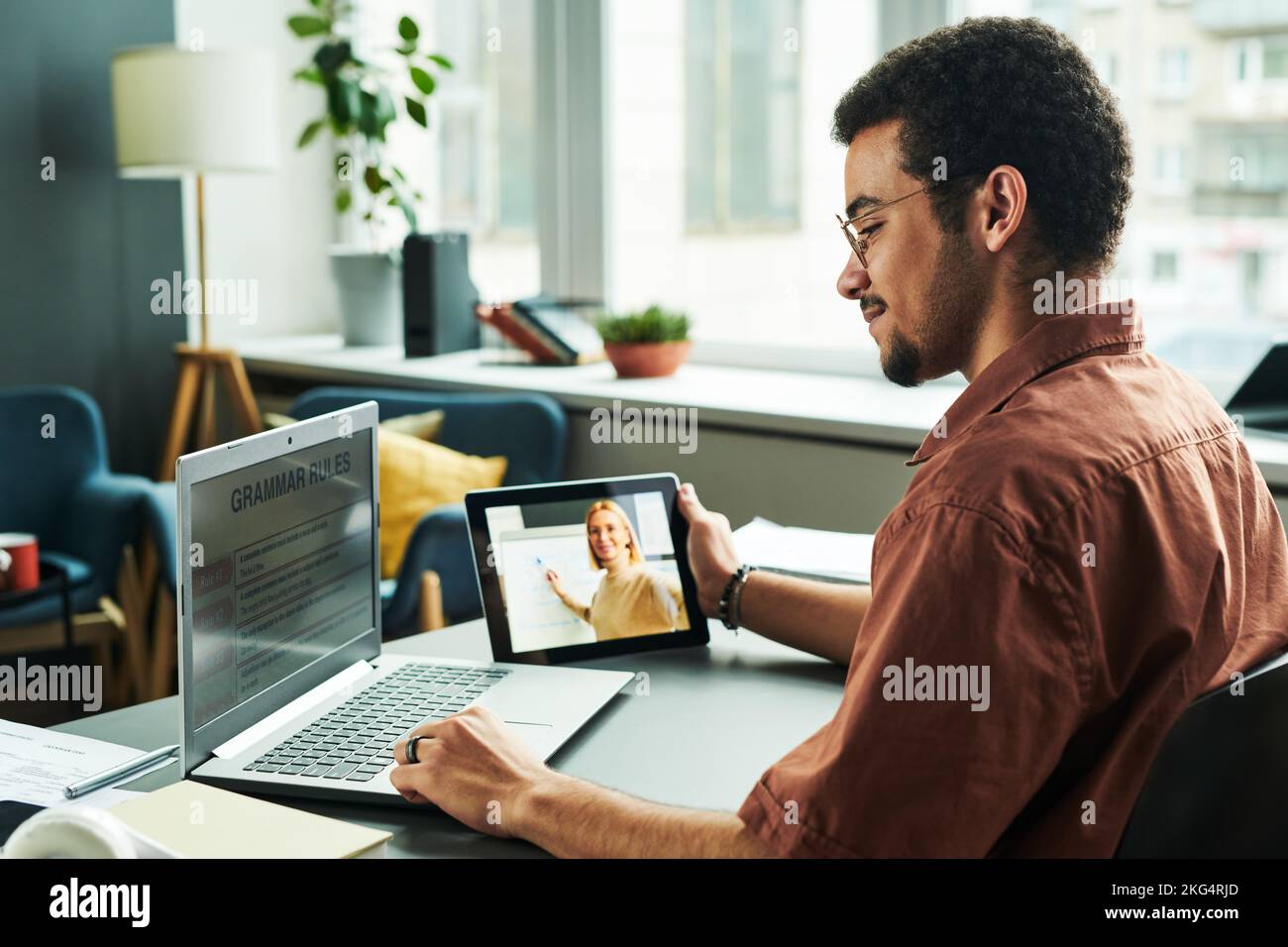 Seitenansicht eines jungen Schülers, der während des Online-Unterrichts Grammatikregeln auf dem Laptop-Bildschirm liest, während er sich die Erklärung des Lehrers auf dem Tablet anhört Stockfoto
