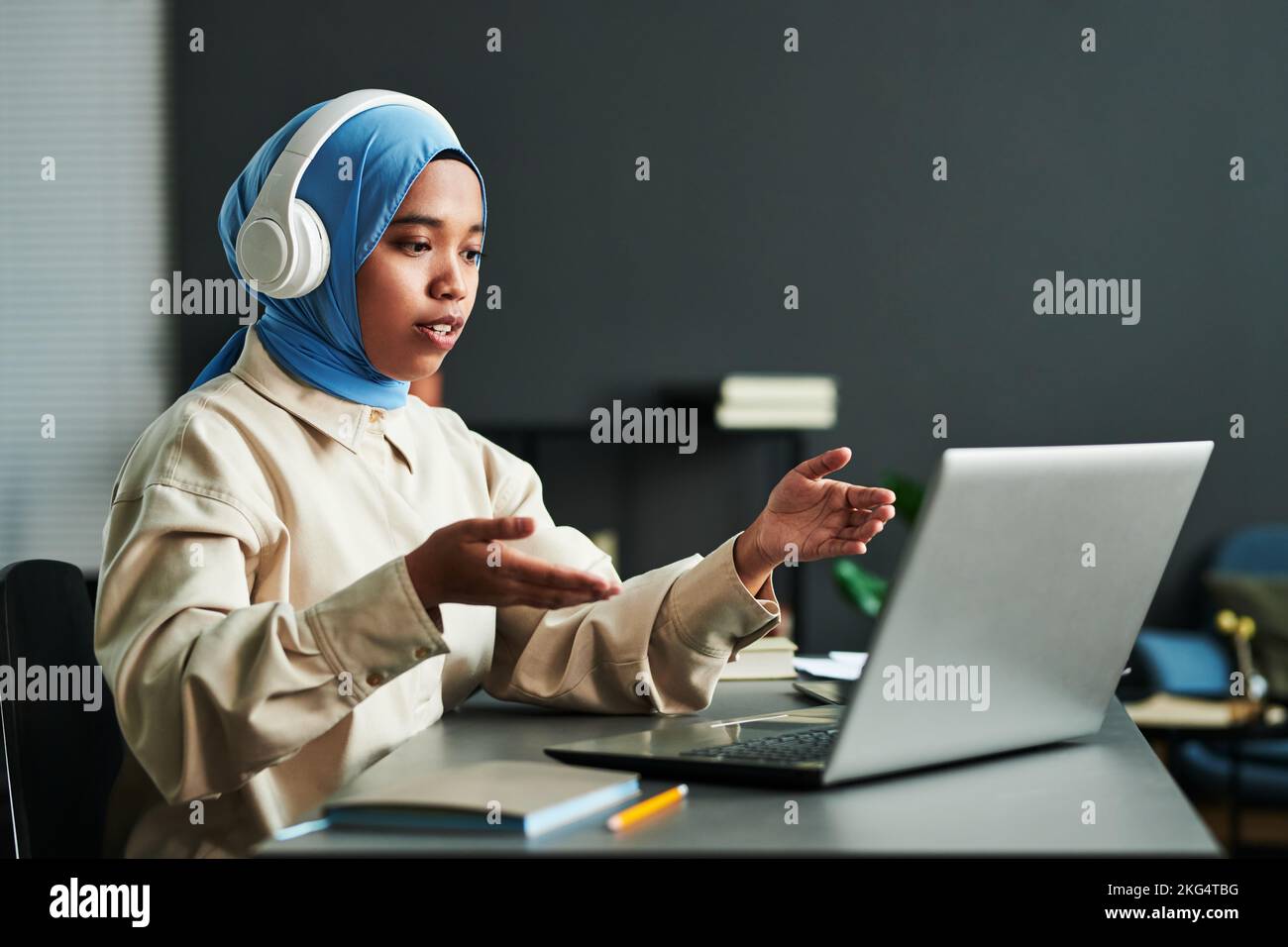 Junge selbstbewusste muslimische Frau in Kopfhörern über blauem Hijab, die während der Online-Kommunikation etwas erklärt Stockfoto