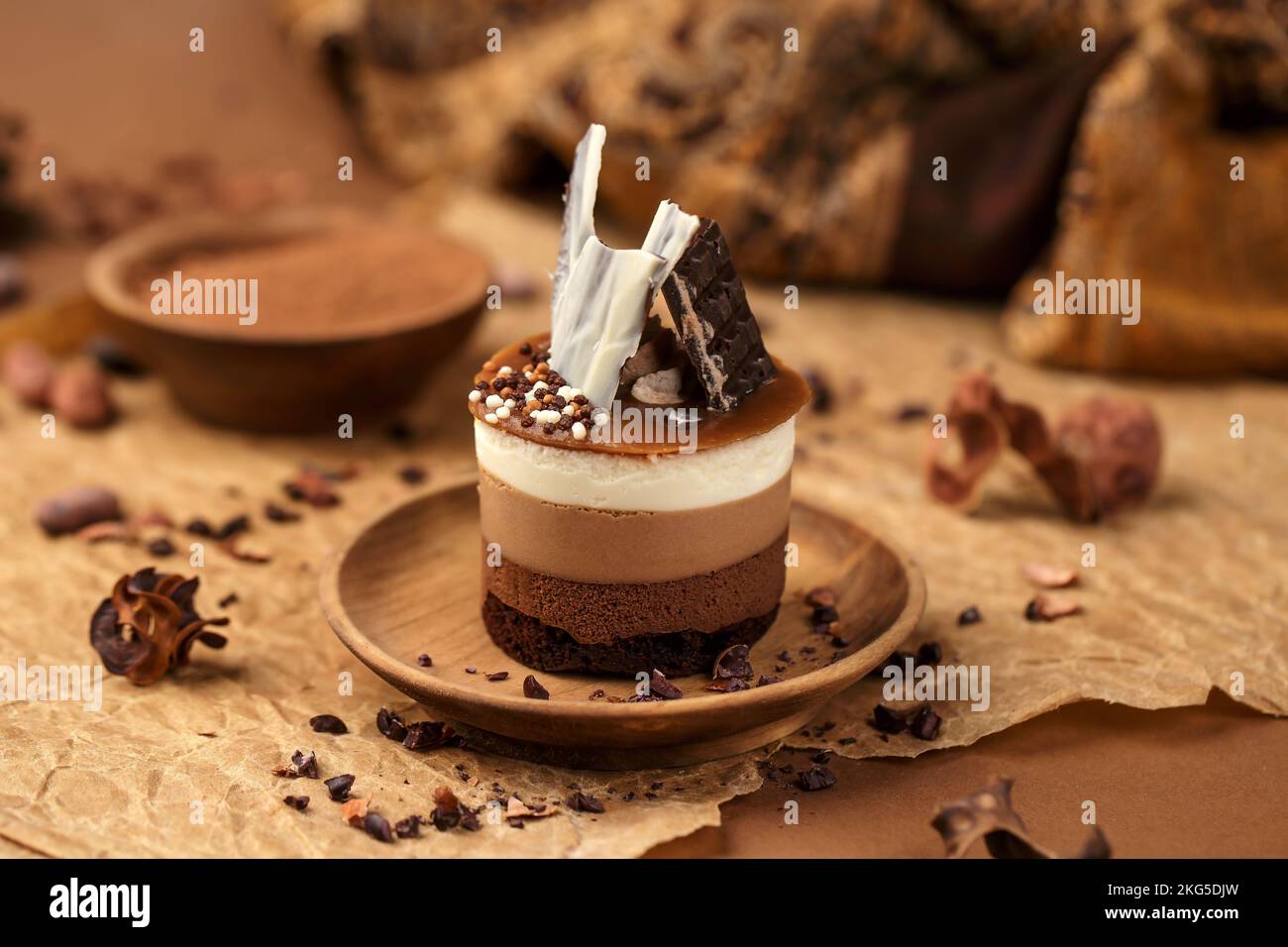 Eine Nahaufnahme von Triple Chocolate Mousse Kuchen, verziert mit einer Schokolade auf einem Holzteller, mit Kakaopulver im Hintergrund Stockfoto