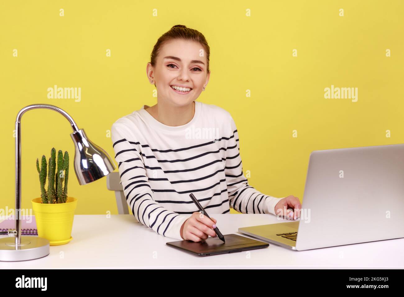 Eine schöne lächelnde kreative Designerin, die am Arbeitsplatz mit einem Tablet und einem Notebook sitzt und mit fröhlichem Ausdruck in die Kamera schaut. Studioaufnahmen im Innenbereich isoliert auf gelbem Hintergrund. Stockfoto