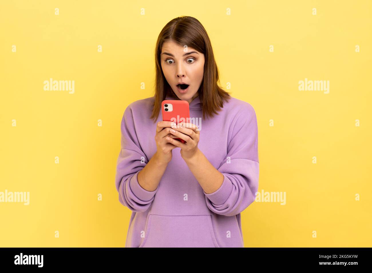 Porträt einer überraschten, aufgeregten Frau, die Nachrichten auf einem Smartphone liest, ein Mobilgerät zur Kommunikation nutzt, von der App überrascht ist und einen lila Hoodie trägt. Innenstudio-Aufnahme isoliert auf gelbem Hintergrund. Stockfoto
