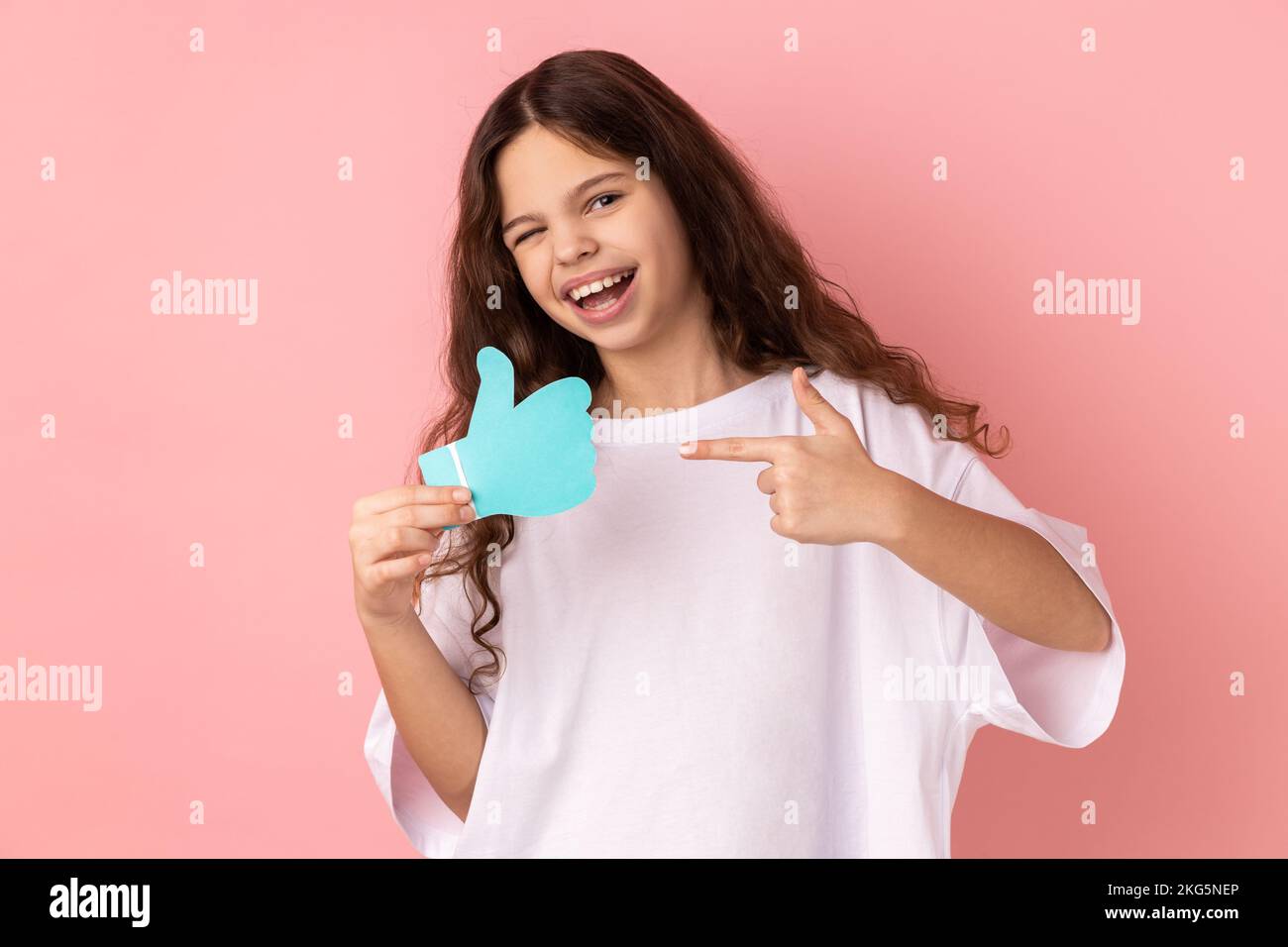 Porträt eines glücklichen, zufriedenen, bezaubernden kleinen Mädchens, das ein weißes T-Shirt trägt und auf ein Schild in Papierform zeigt oder Daumen nach oben zeigt. Studio-Aufnahme im Innenbereich isoliert auf pinkfarbenem Hintergrund. Stockfoto