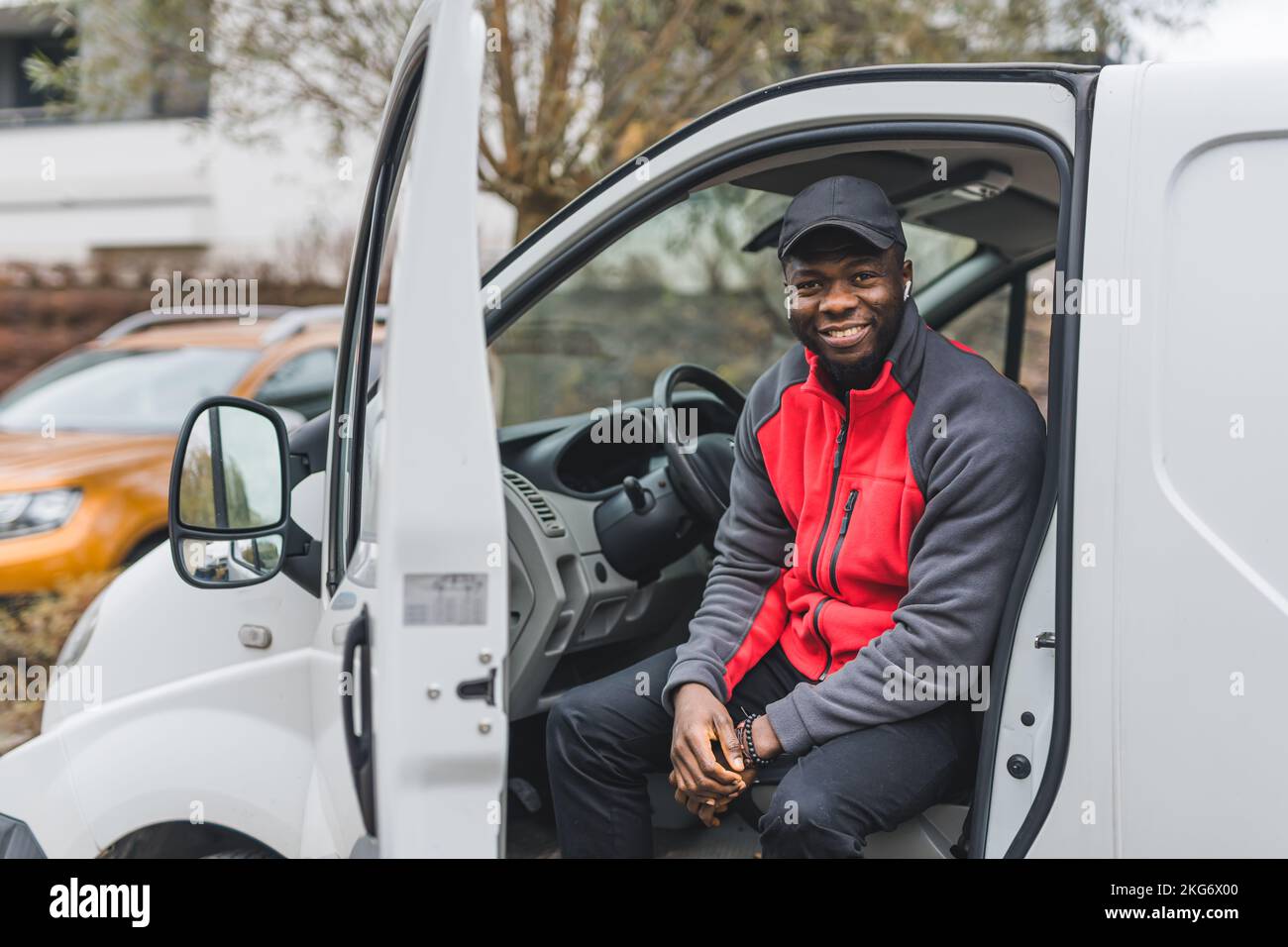 Beruf - Auslieferer. Positiv lächelnder Schwarzer 25-jähriger Mann in Postzustelluniform in rot und schwarz auf dem Fahrersitz. Hochwertiges Foto Stockfoto
