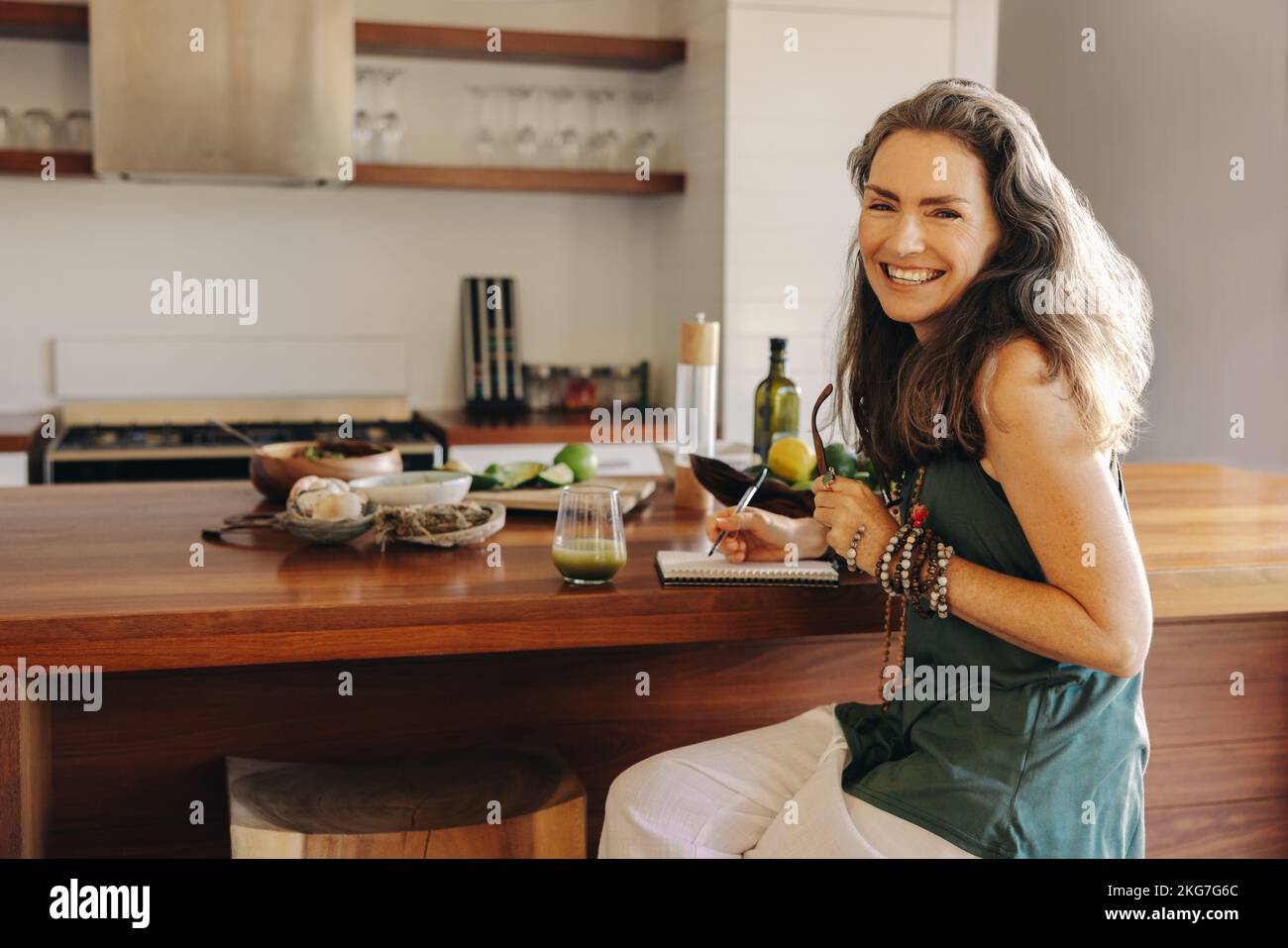 Eine vegetarische Frau lächelt in die Kamera, während sie ihre Essenspläne und Rezepte aufschreibt. Reife Frau plant eine gesunde pflanzliche Ernährung in ihrem Tagebuch. H Stockfoto