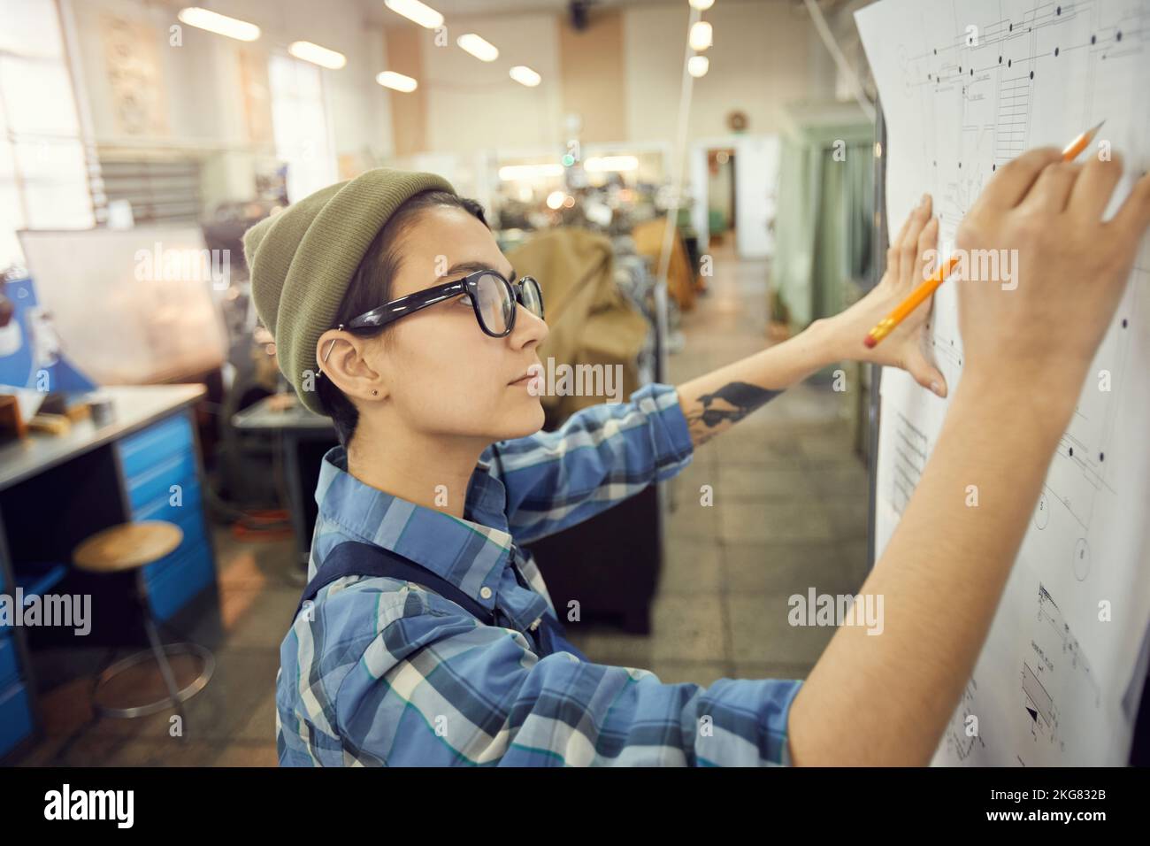 Seitenansicht einer fokussierten jungen Frau in einer Brille, die im Fabrikgebäude steht und technische Skizzen an die Wand lehnt Stockfoto