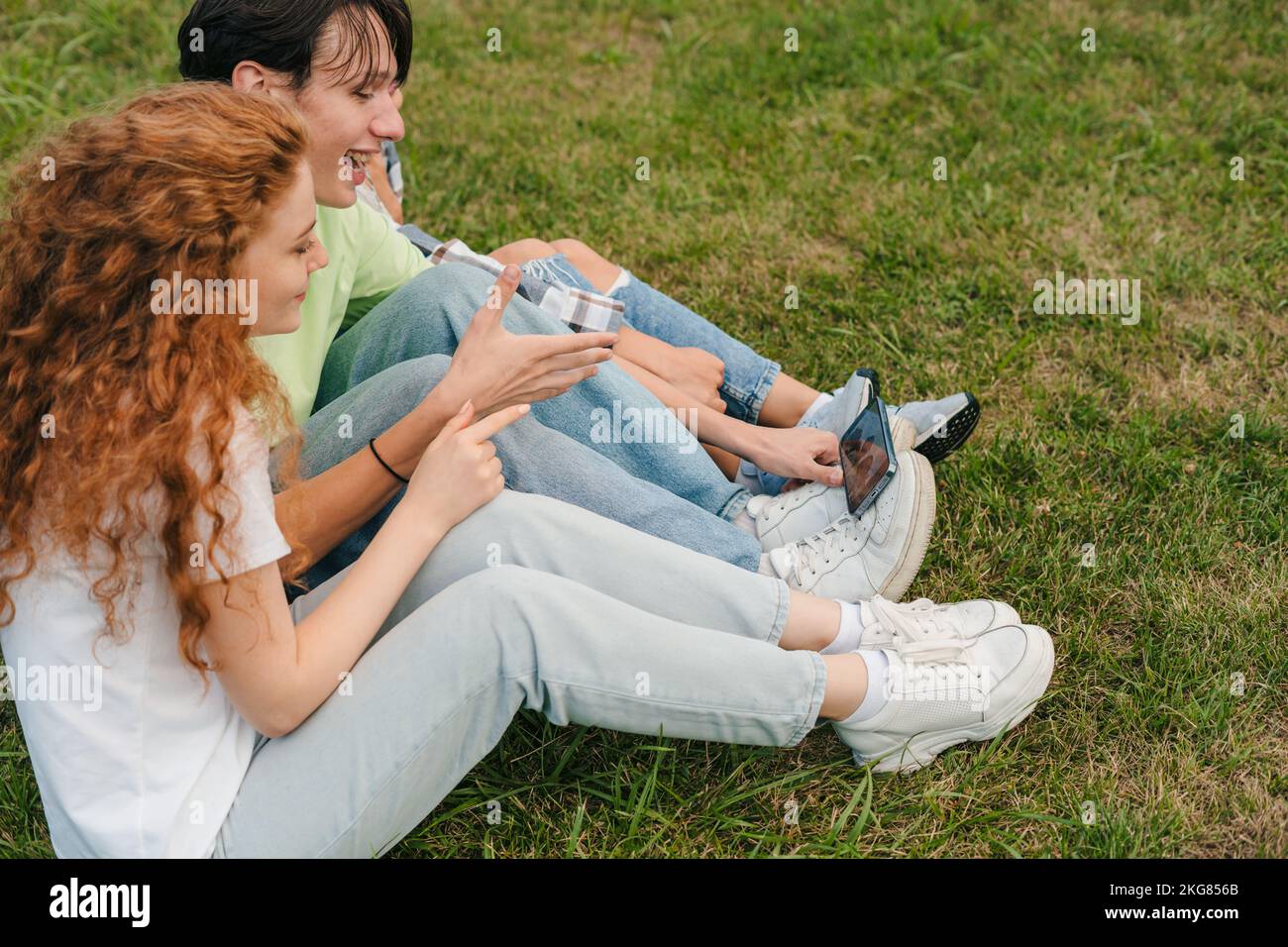 Unbeschwerte glückliche Freunde, die Video-Blog aufzeichnen, auf dem Smartphone-Bildschirm sitzend, auf grünem Gras im Park im Freien. Portrait für Lifestyle-Design. Mobil Stockfoto