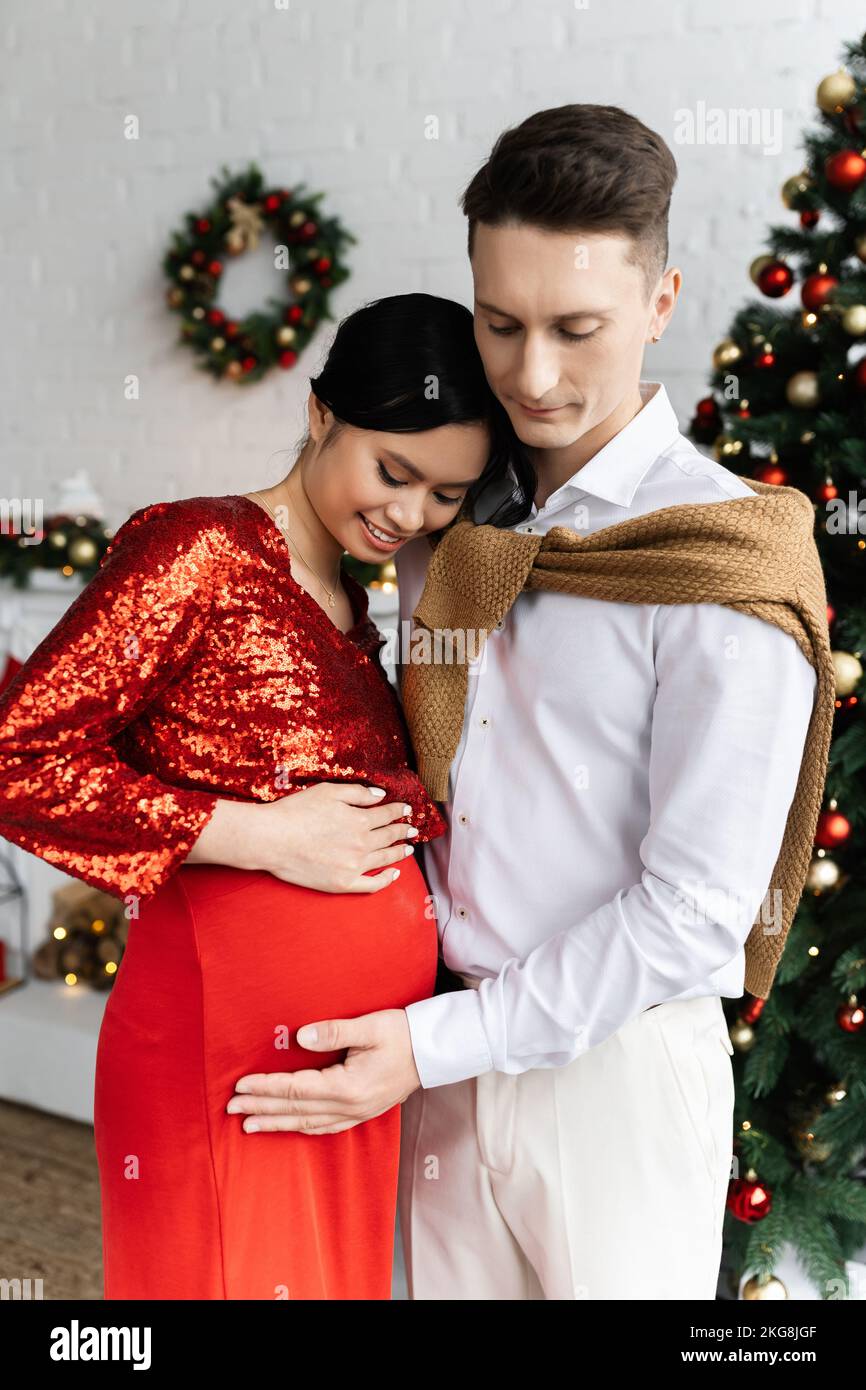 Mann berührt den Bauch einer lächelnden und schwanger asiatischen Frau in festlicher Kleidung in der Nähe verschwommenes Weihnachtsdekor Stockfoto