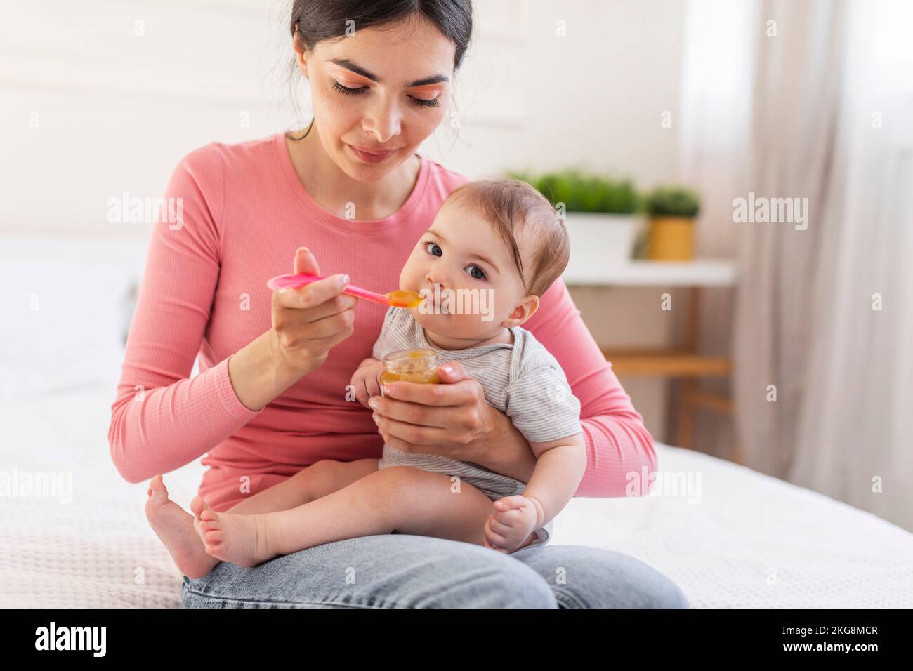 Ein bezauberndes kleines Mädchen, das aus dem Löffel isst, eine junge fürsorgliche Mutter, die ein süßes Baby mit Fruchtpüree füttert Stockfoto