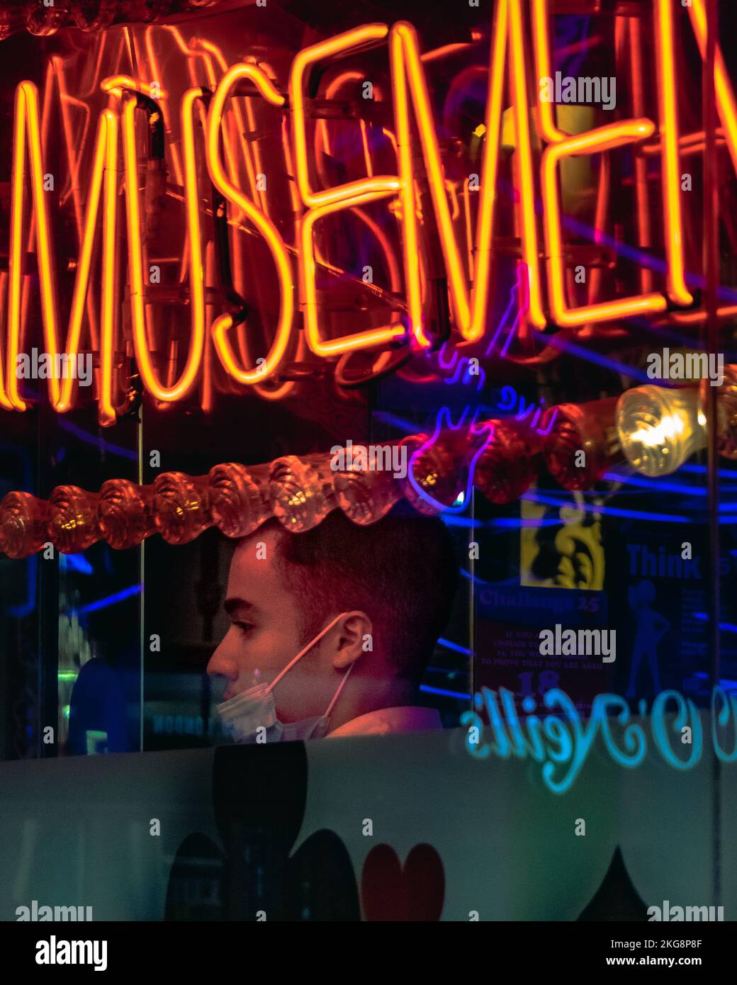 Ein Mann mit einer Gesichtsmaske unter dem Kinn in einer Spielhalle in Chinatown, soho, London, eingerahmt von einem Neonschild im Fenster. Stockfoto