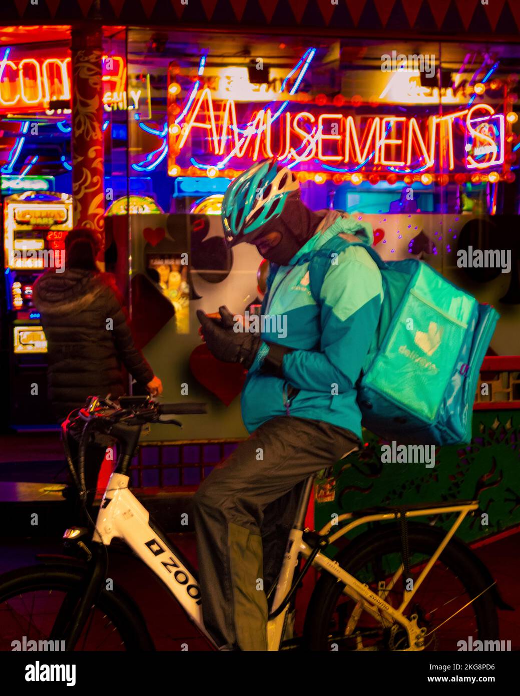 Ein Mann in einer Auslieferungsuniform, der eine Umhängetasche in einem Fahrradhelm trug, Fahrrad fuhr und sein Handy in London anschaute. Stockfoto