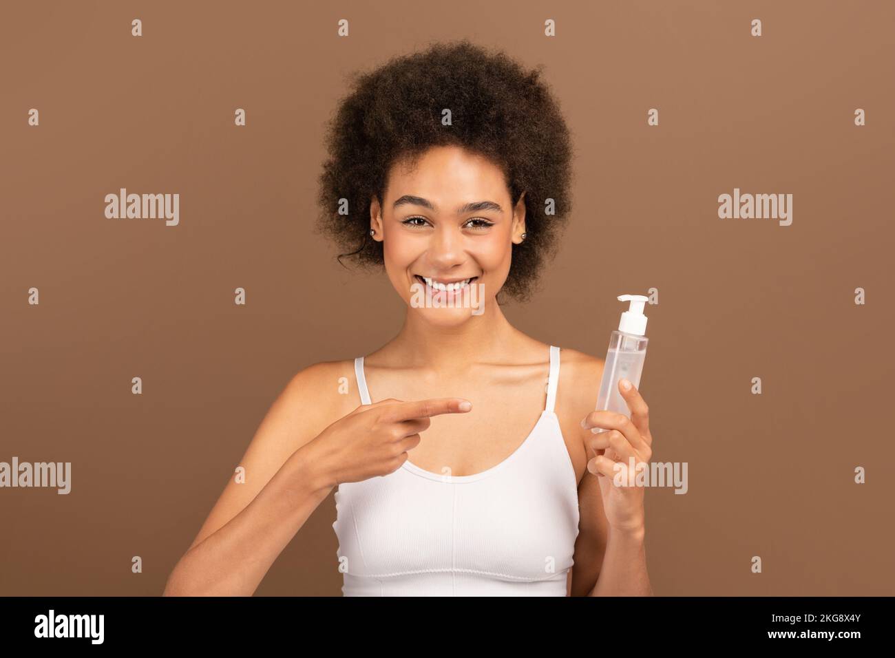 Fröhliche, afroamerikanische, lockige Dame mit weißem, oberem Finger, der auf ein Glas Lotion für Haare oder Haut zeigt Stockfoto