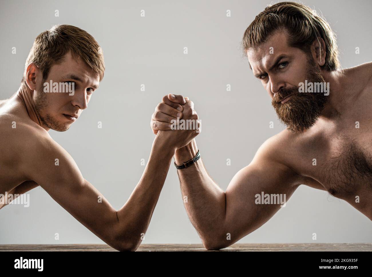 Armdrücken. Stark muskulös bärtigen Mann Arm Ringen ein mickrigen schwachen Mann. Arme ringen dünne Hand, großer starker Arm im Studio. Zwei Männerhände umklammten Stockfoto