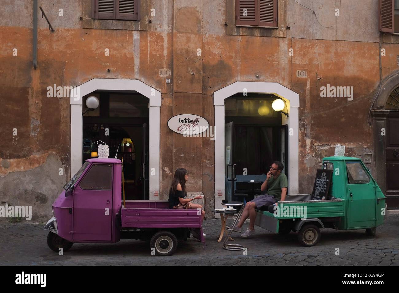 Rom, Italien: Der Mann sitzt auf dem Rücksitz eines offenen grünen Lkws und spricht mit einer Frau, die auf einem kleinen pinken Truck chillt. Fahrzeuge parken vor dem Lettere Caffe in Trastevere. Stockfoto