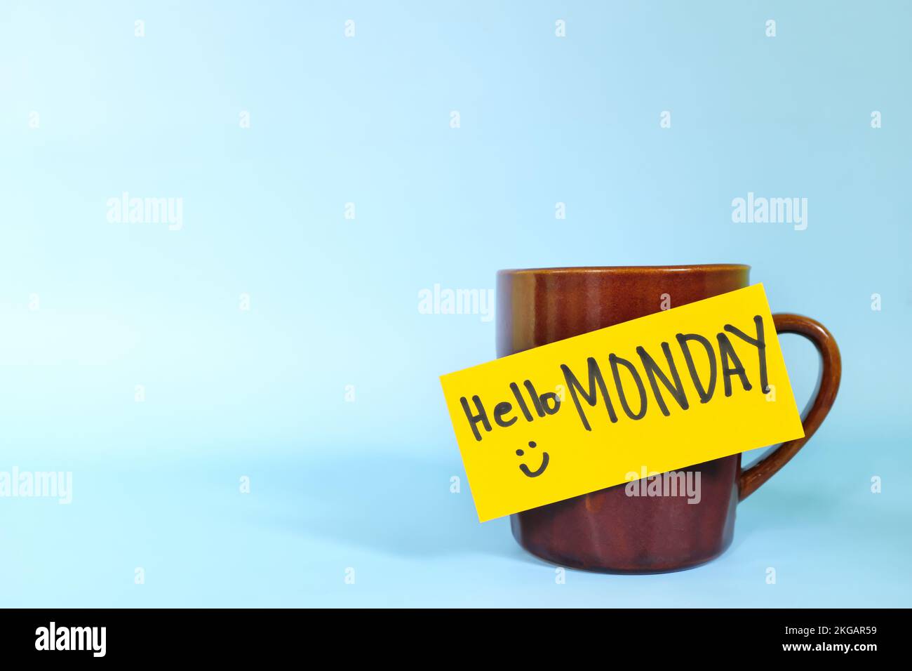 Willkommen, hallo und Happy Monday Concept. Selektiver Fokus der Kaffeetasse mit hellgelbem Papierzettel und schriftlicher Nachricht isoliert auf blauem Hintergrund. Stockfoto
