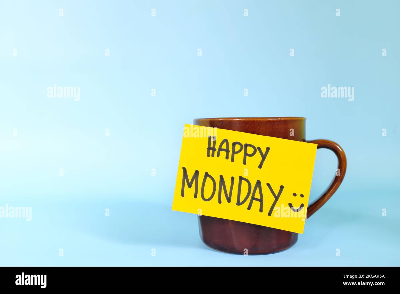 Willkommen, hallo und Happy Monday Concept. Selektiver Fokus der Kaffeetasse mit hellgelbem Papierzettel und schriftlicher Nachricht isoliert auf blauem Hintergrund. Stockfoto