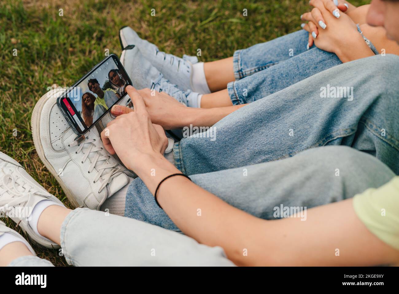 Drei Freunde sitzen auf dem Rasen in einem Park und Filmen sich mit einem Mobiltelefon, das von ihren Schuhen getragen wird. Mobiltelefonkommunikation. Internet Stockfoto