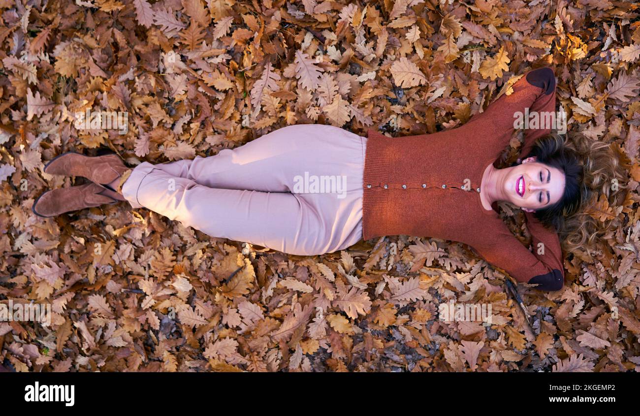 Junge, übergroße, wunderschöne Frau, die im Herbst auf einem Bett mit heruntergefallenen Blättern im Eichenwald liegt Stockfoto