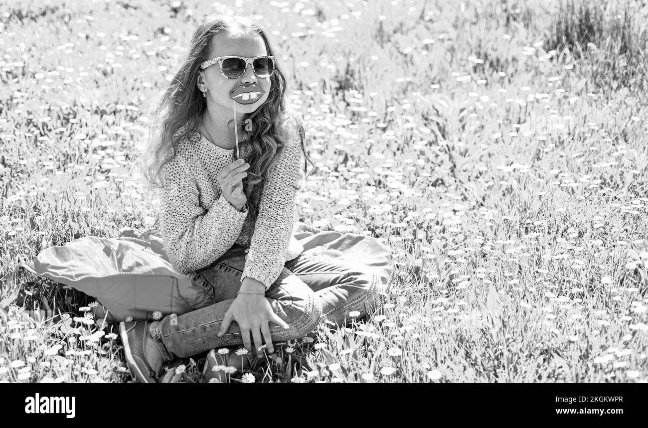 Das Mädchen sitzt auf Gras am Grassplot, grüner Hintergrund. Kinder, die mit Brillen posieren, lächelnde Lippen aus Pappe für eine Fotosession auf der Wiese. Fashionista Stockfoto