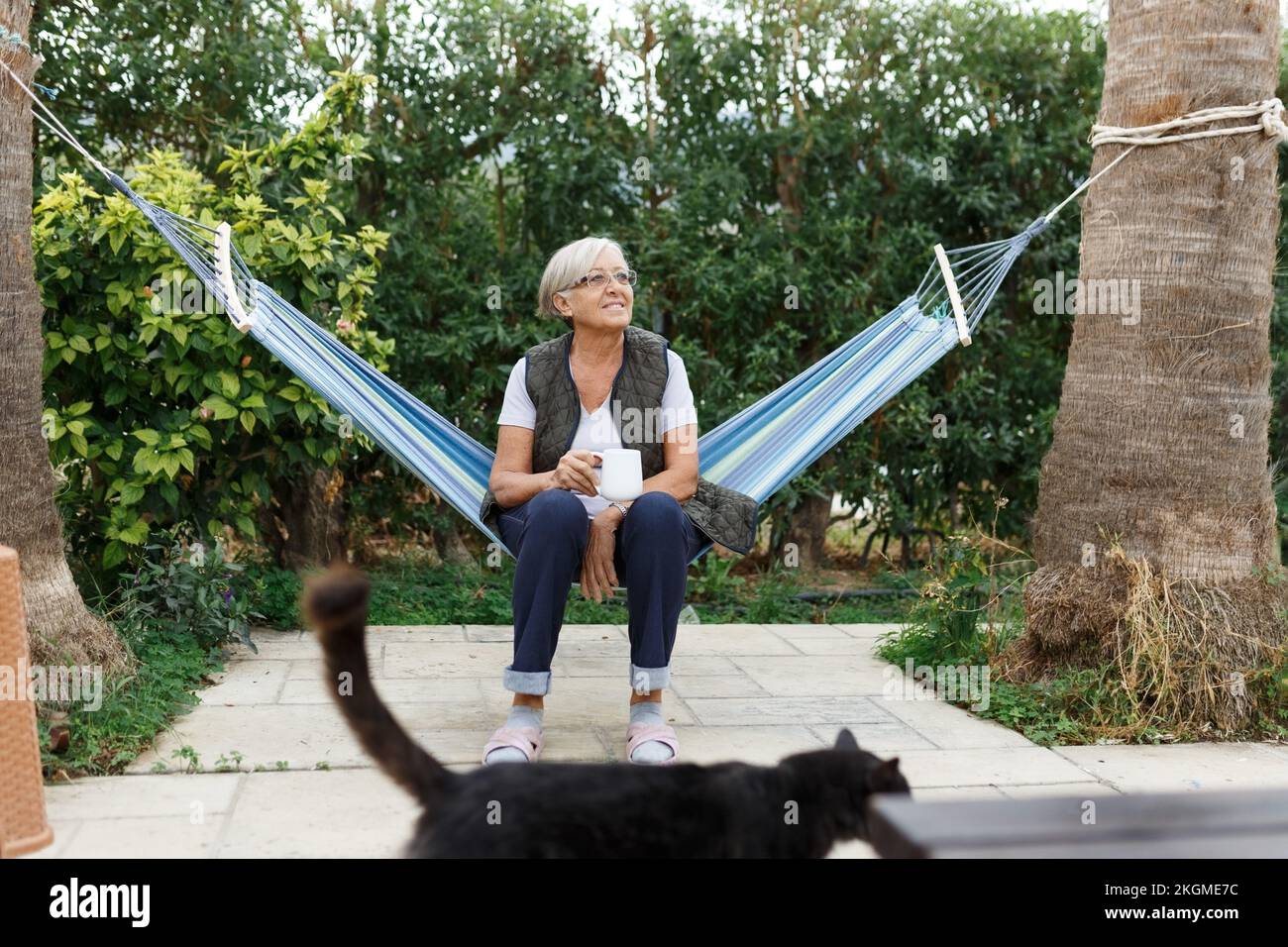 Wunderschöne ältere blonde Frau, die ein Buch liest und mit einer Katze in einer Hängematte im Garten sitzt Stockfoto