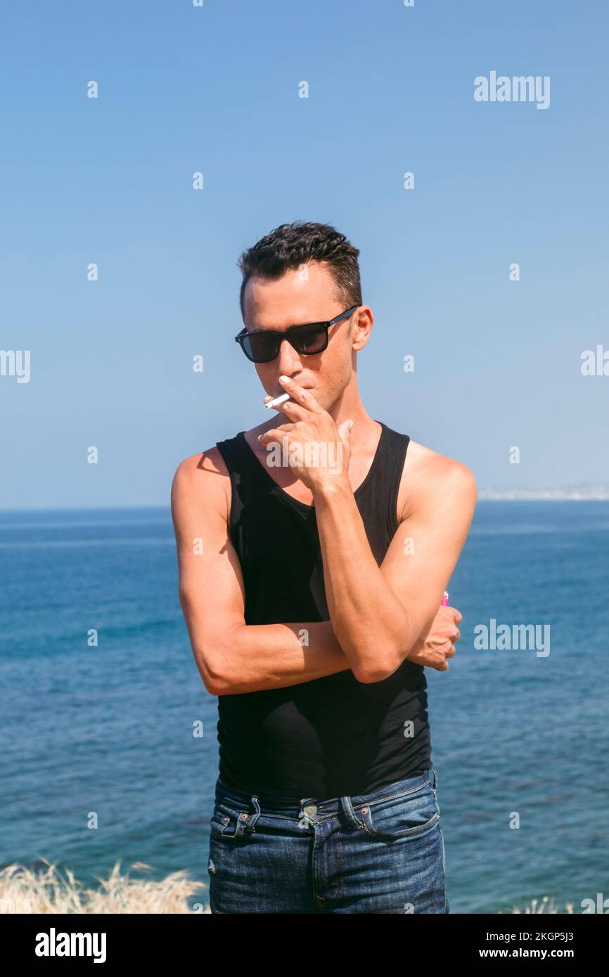 Ein Mann mit Sonnenbrille, der vor dem Meer raucht Stockfoto