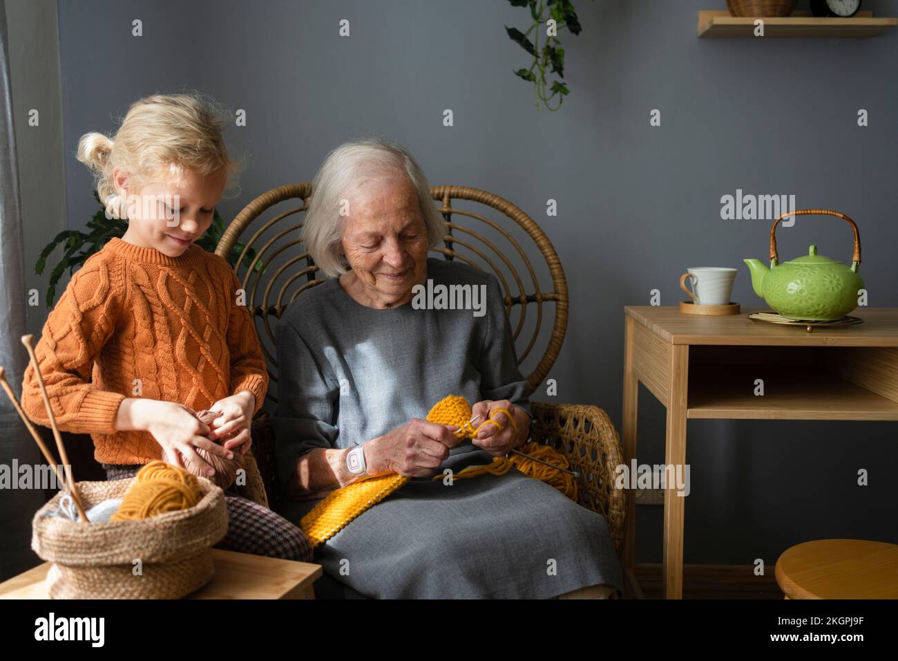 Lächelndes Mädchen, das Wollknäuel hält, steht bei Großmutter und strickt zu Hause auf einem Stuhl Stockfoto