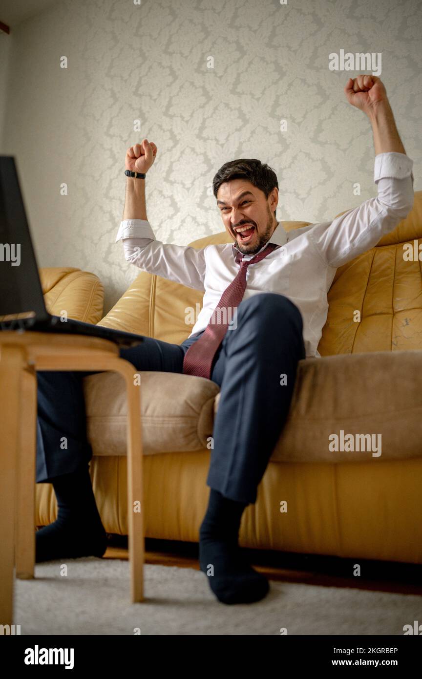 Ein fröhlicher Geschäftsmann, der zu Hause auf dem Sofa sitzt und seine Fäuste streckt Stockfoto