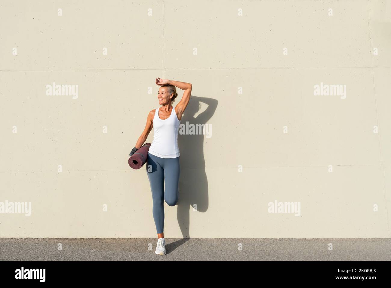 Eine Frau mit Yoga-Matte, die sich an die cremefarbene Wand lehnt Stockfoto