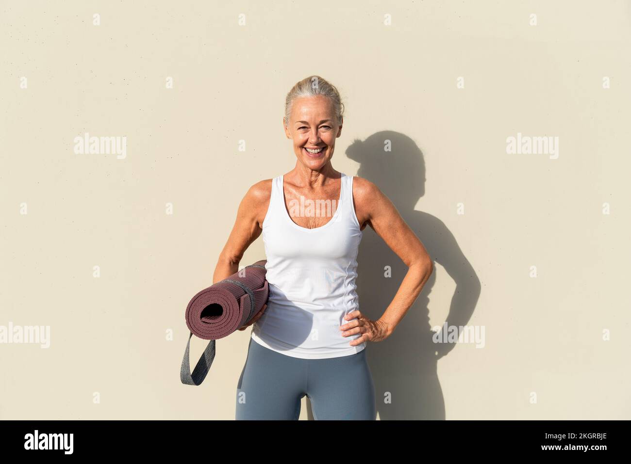 Lächelnde Frau mit Yogamatte vor cremefarbenen Wänden Stockfoto