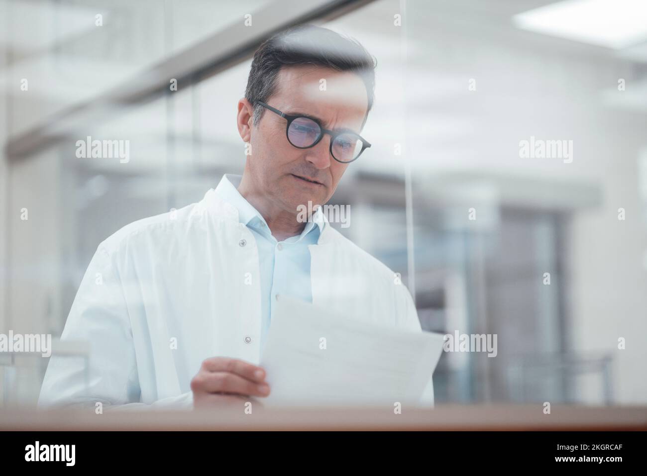 Mann im Laborkittel analysiert medizinische Berichte im Labor, durch Glas gesehen Stockfoto