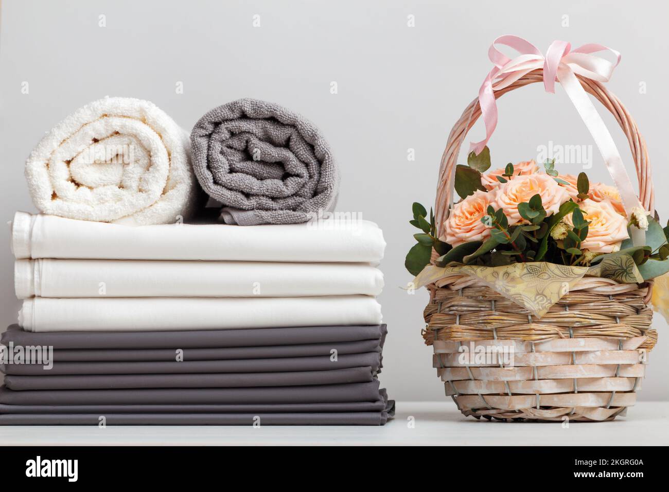 Ein Stapel Bettwäsche, Rollen mit weißen und grauen Handtüchern. Korb mit Rosen auf dem Tisch. Stockfoto