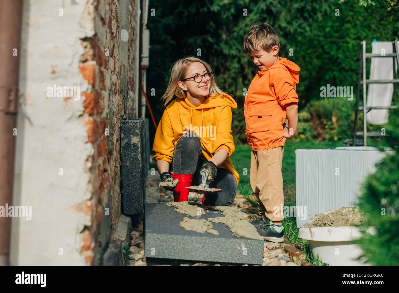 Lächelnde Mutter, die Zement auf Polystyrol streut und den Sohn im Hof sieht Stockfoto