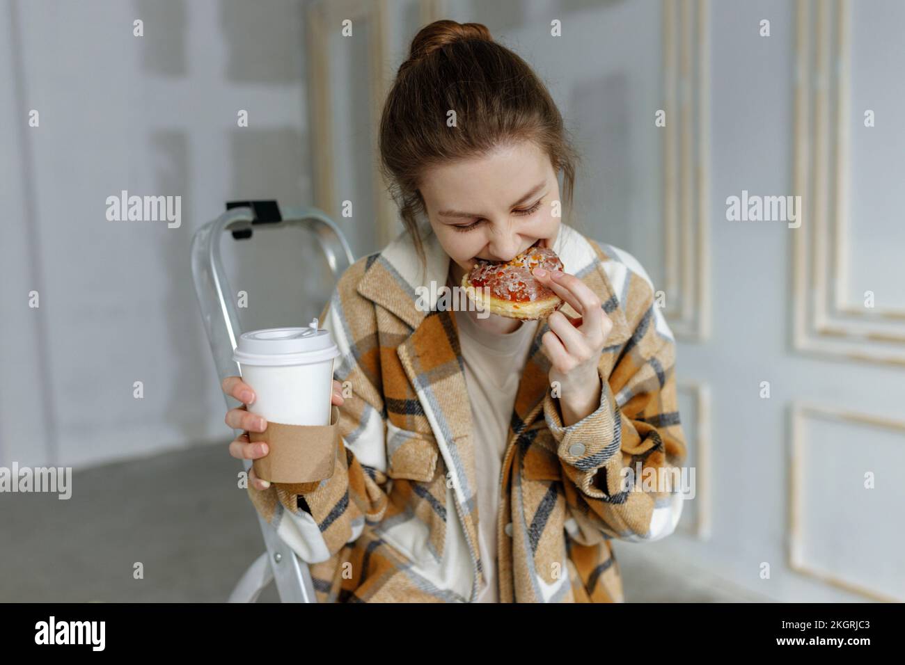 Junge Frau mit Einweg-Kaffeetasse, die in der Wohnung Essen isst Stockfoto