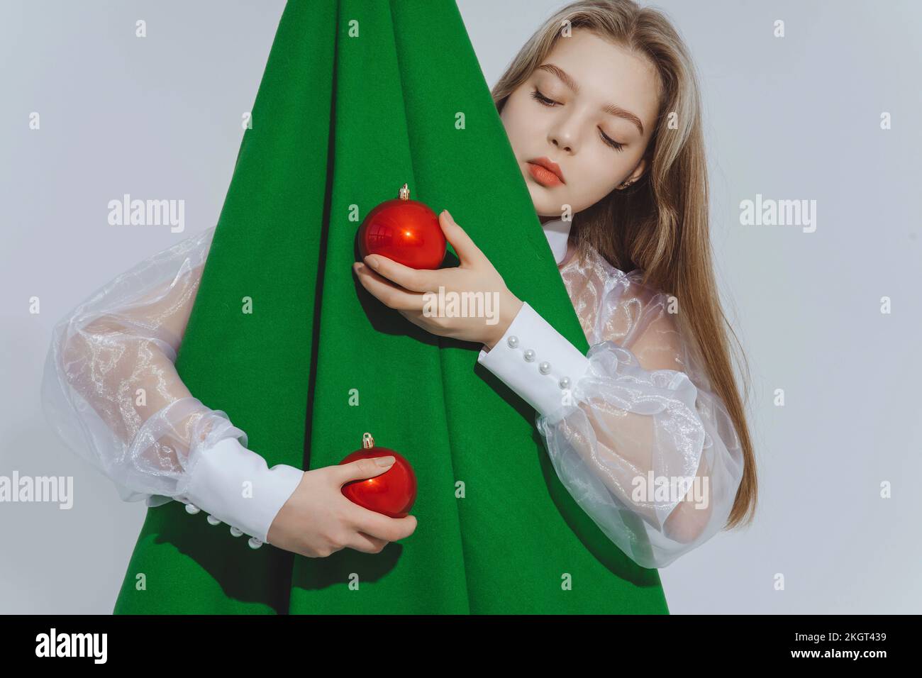 Ein Mädchen mit roten Kugeln, das einen abstrakten Weihnachtsbaum vor weißem Hintergrund umarmt Stockfoto