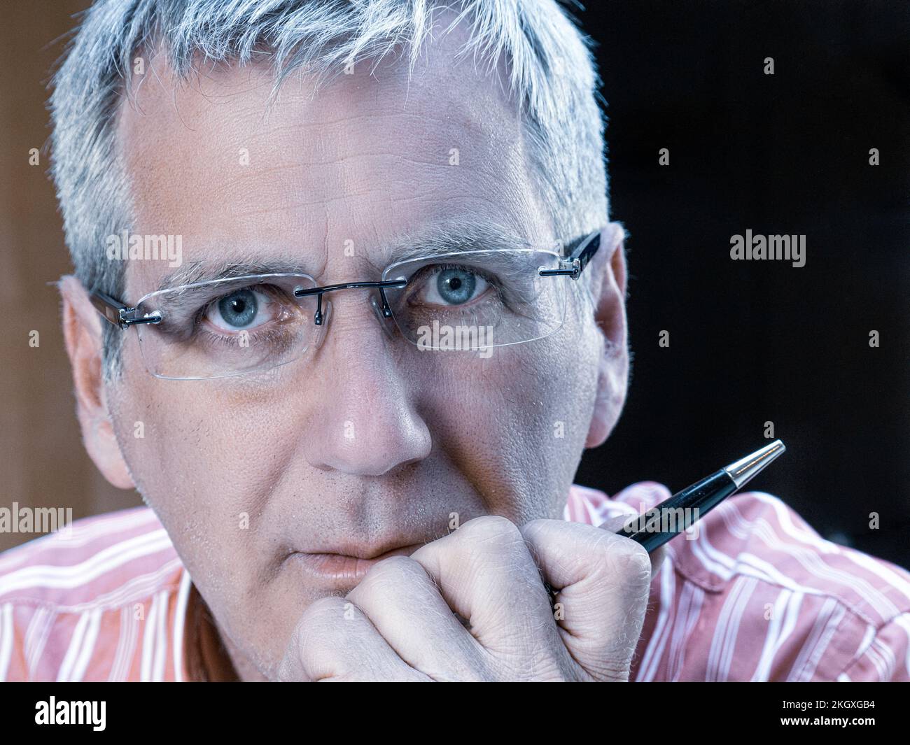 Geschäftsmann männlich 50s-Gesicht, reifer Brillenträger, Hand am Kinn hält einen Stift, der direkt in die Kamera schaut, mit ernstem Gesichtsausdruck Stockfoto
