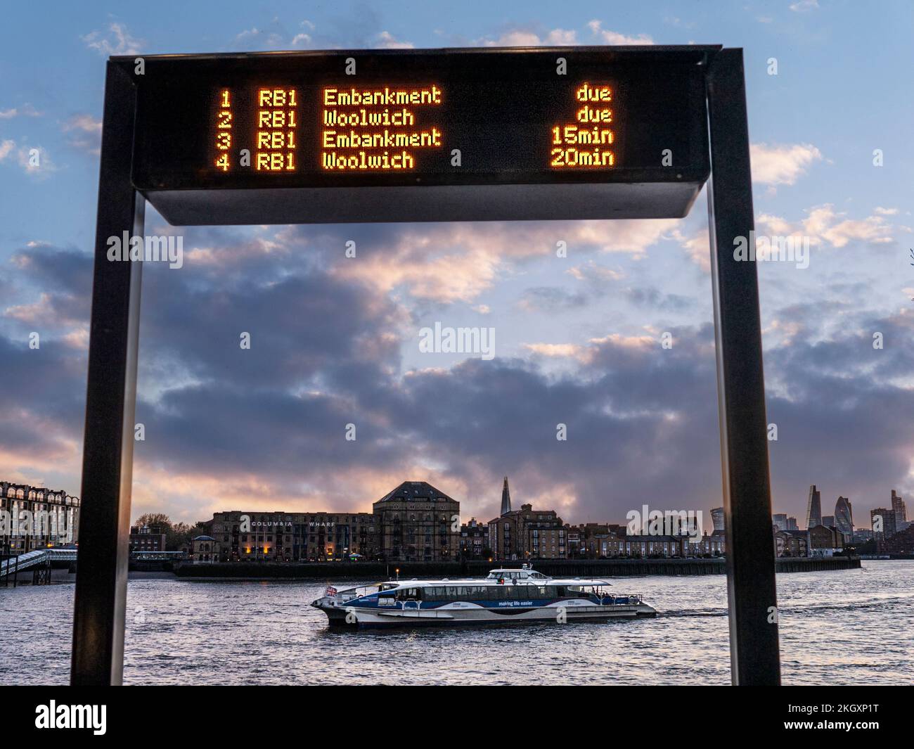 CANARY WHARF CLIPPER RIVER BOAT RB1 Digital Arrivals Information Screen und RB1 Thames Clipper River Boat Arrival at Sunset Arrival in Canary Wharf London UK mit City of London einschließlich The Shard Behind. Stockfoto