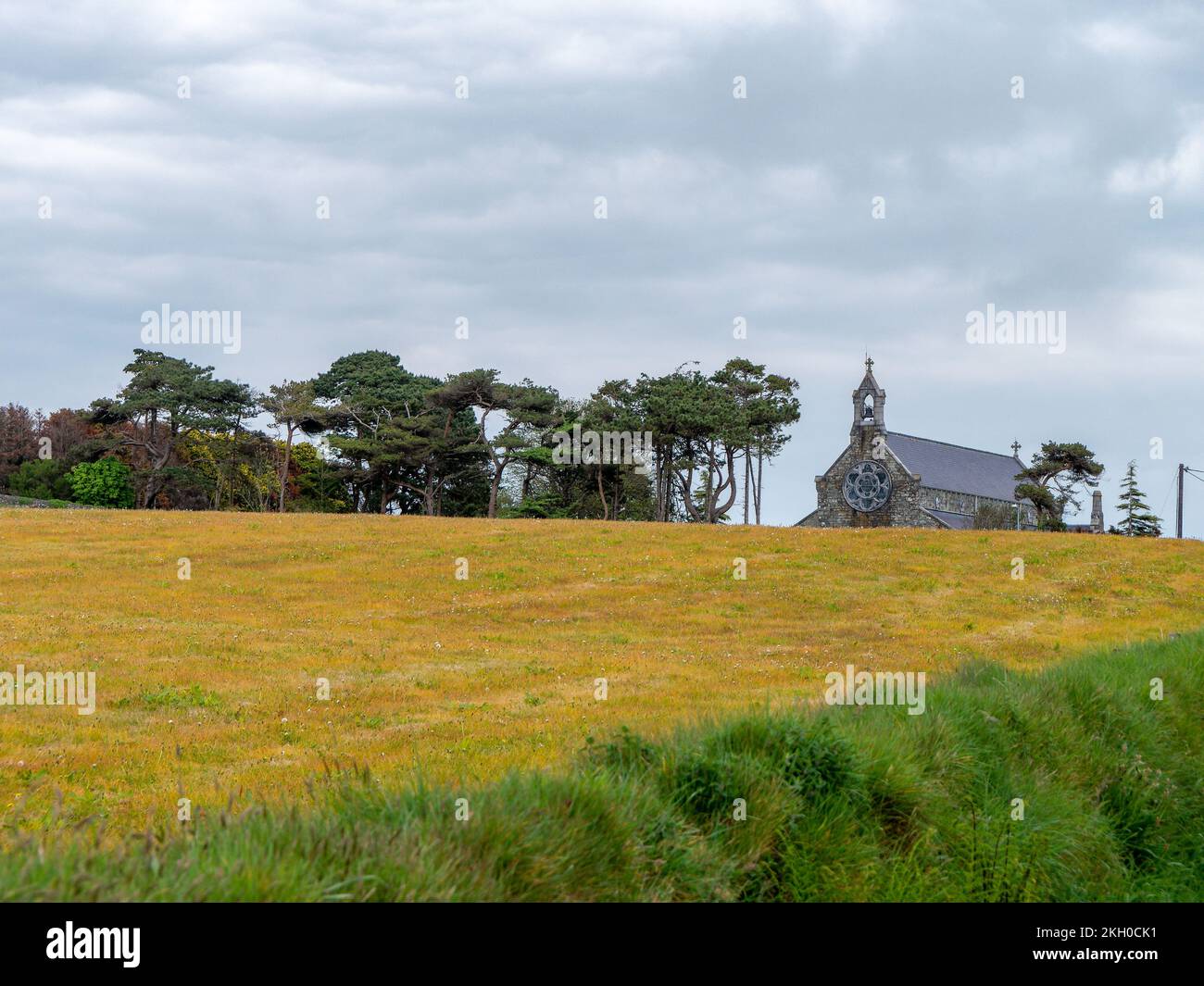 Eine kleine christliche Steinkirche in der Ferne einer wunderschönen Wiese. Wolkiger Himmel, europäische Landschaft. Grün-gelbes Grasfeld mit Bäumen unter W. Stockfoto Eine kleine christliche Steinkirche in der Ferne einer wunderschönen Wiese. Wolkiger Himmel, europäische Landschaft. Grün-gelbes Grasfeld mit Bäumen unter W. Stockfoto