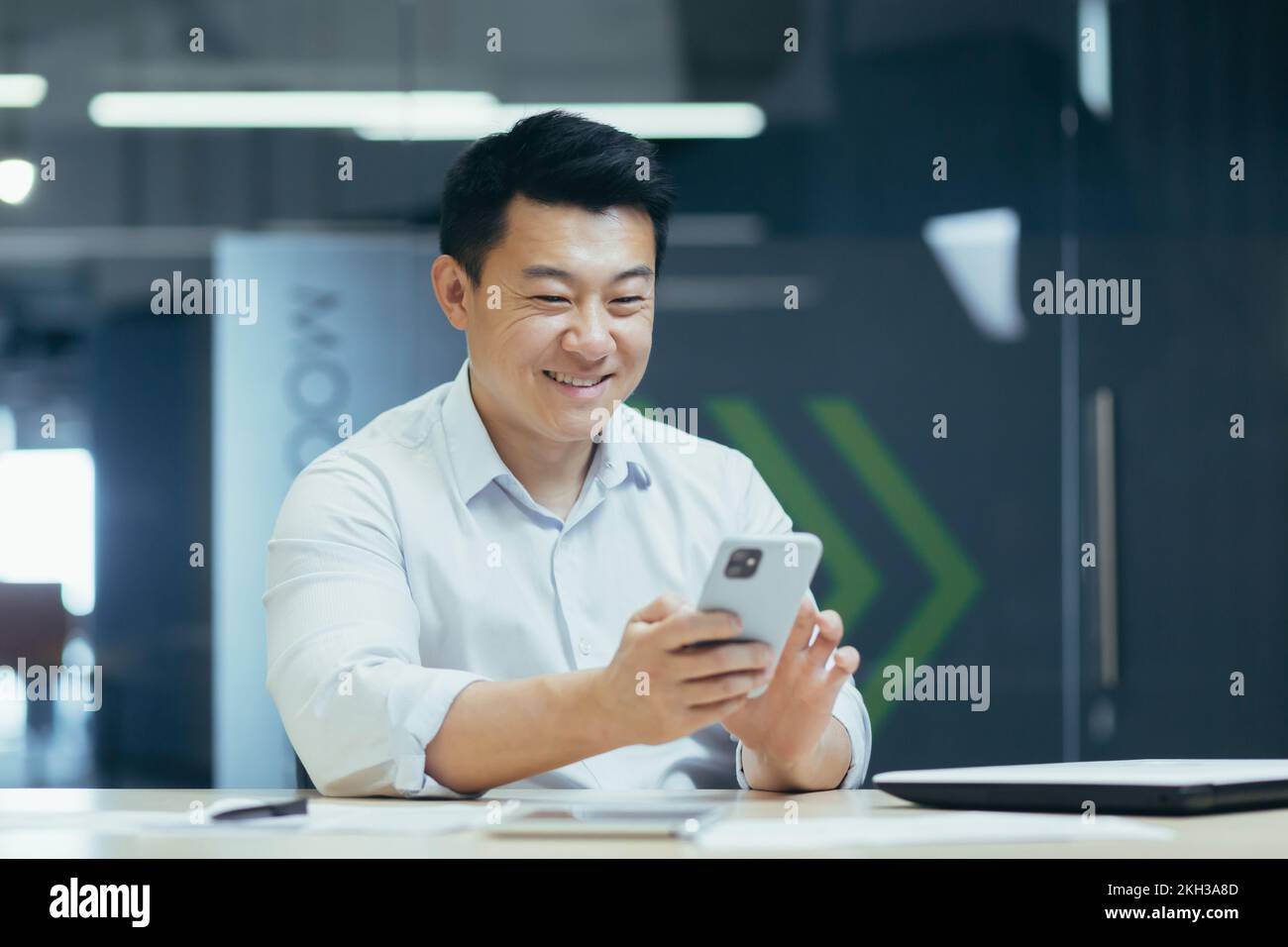 Junger, gutaussehender Asiate, Büroangestellter sitzt am Tisch im Büro, spielt mit dem Telefon, tippt Nachrichten, chattet. Stockfoto